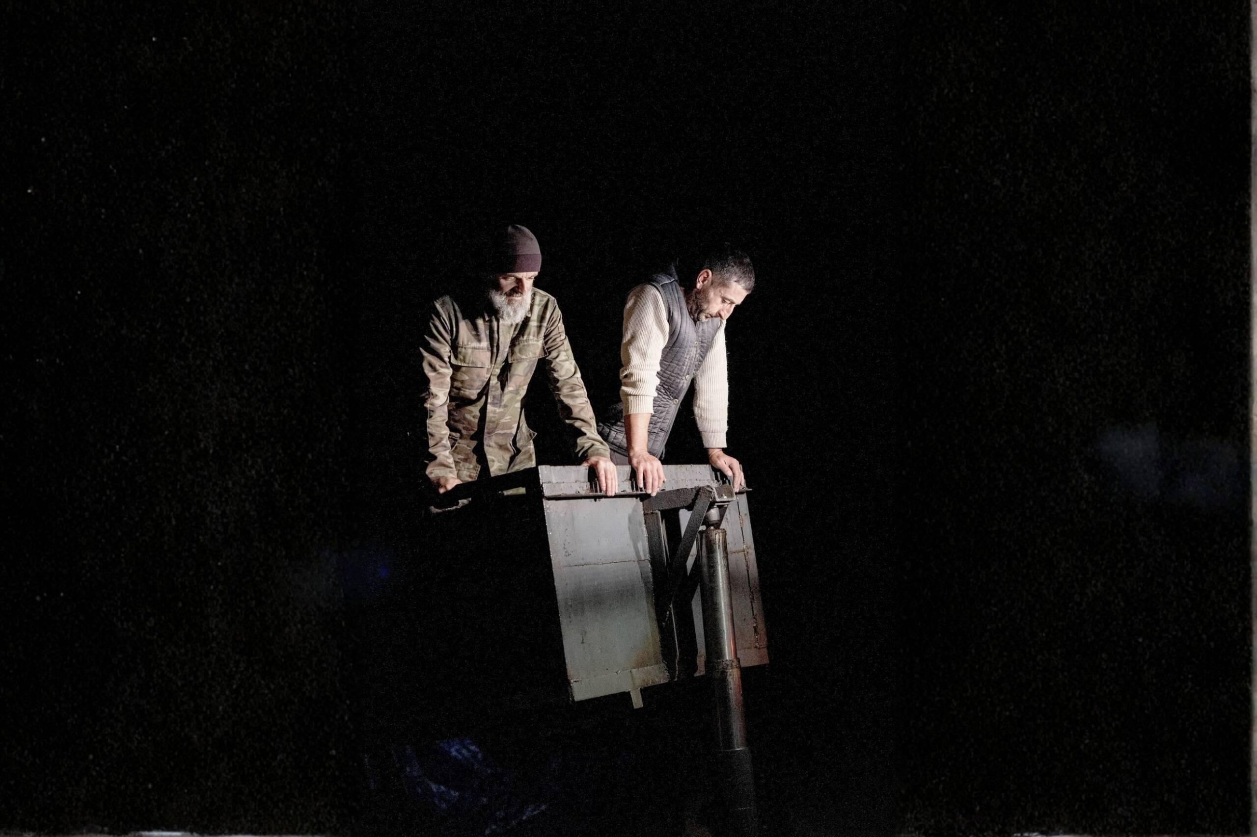 Two men stand on top of a metal container at night, illuminated by a single light source in an otherwise dark vineyard in Kakheti, Georgia.
