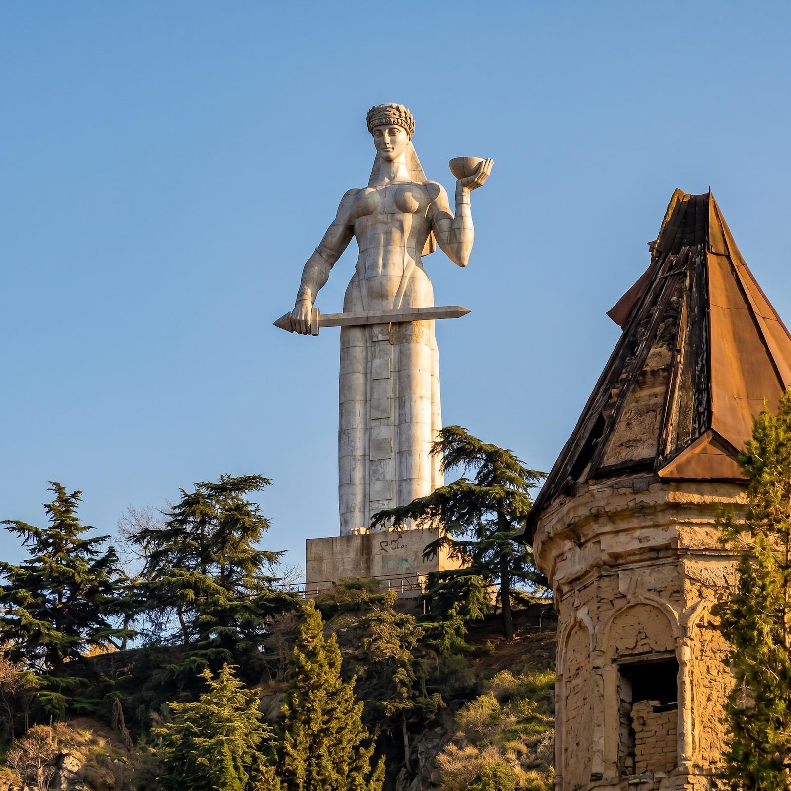 A tall gleaming statue of "The Mother of Georgia" holding a sword and a bowl stands on a hill surrounded by trees, with part of an ancient tower in the foreground.