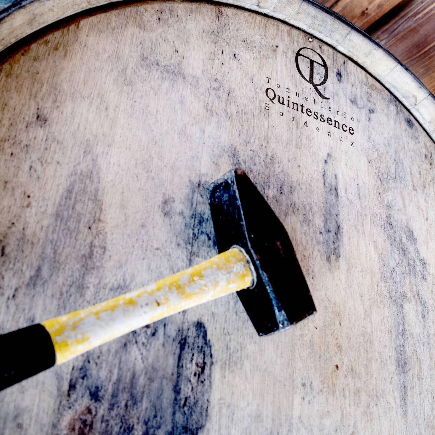 A black and yellow hammer rests on the wooden top of a wine barrel labeled "Tonnellerie Quintessence Bordeaux."