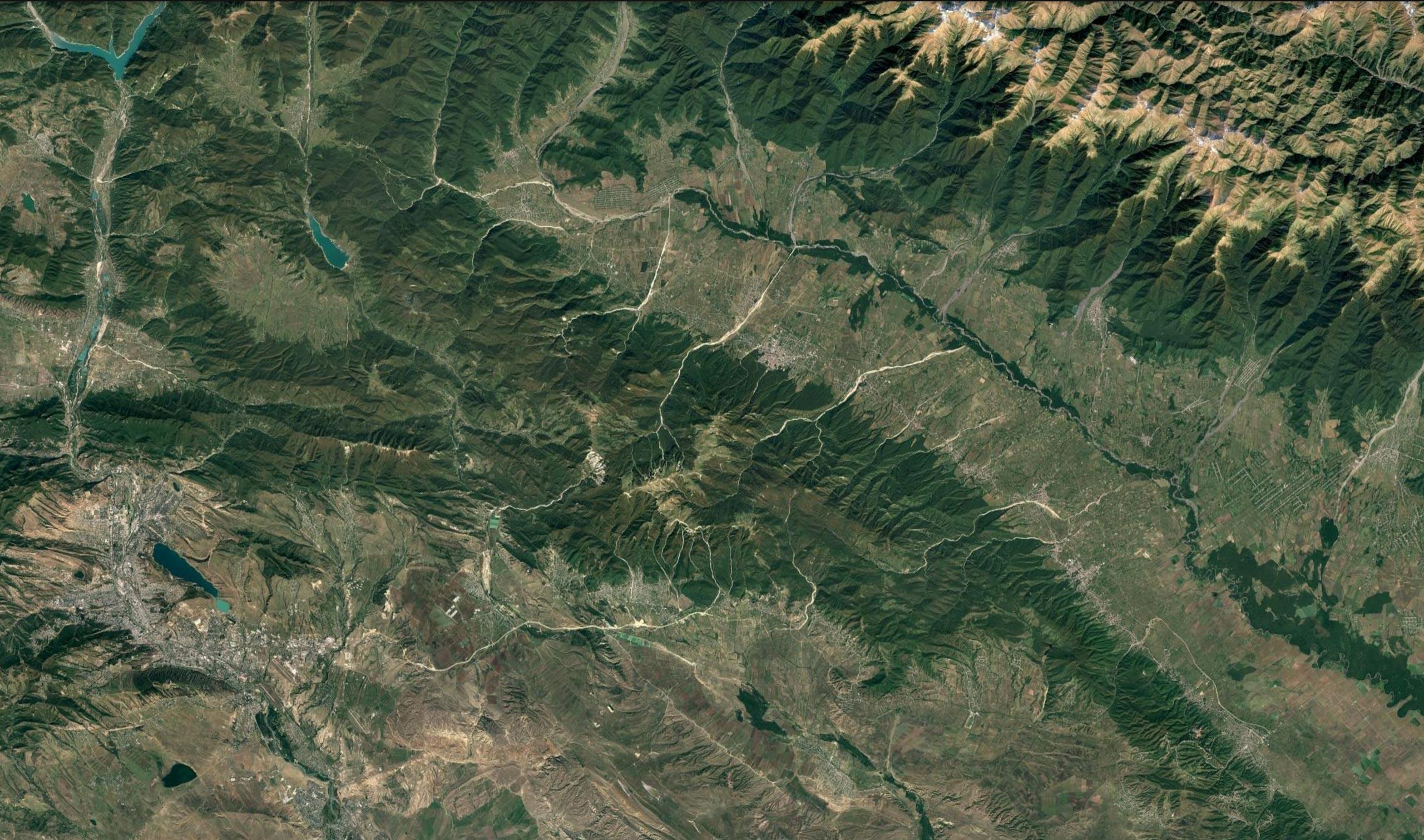 A satellite view of a mountainous region with visible ridges, valleys, rivers, and a city area in the lower left corner.
