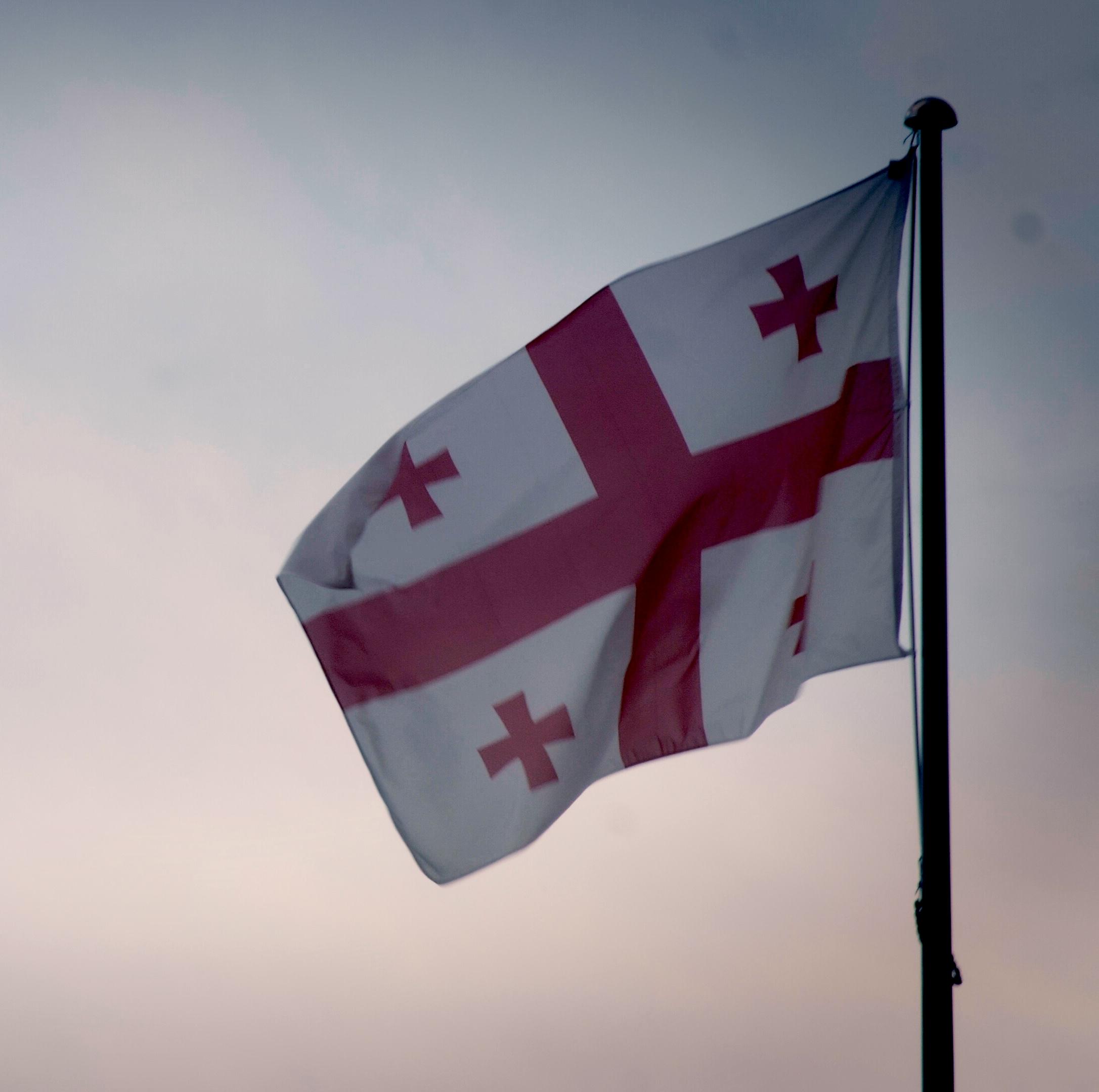 The flag of Georgia with a large red cross and four smaller crosses on a white background, flying on a flagpole against a cloudy sky.