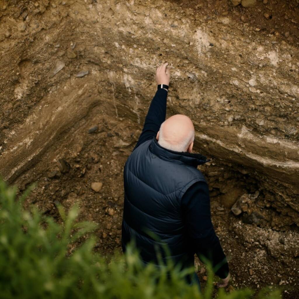 Irakli Gilauri, in a black vest, stands in a deep dirt hole, reaching up to touch the earth wall. Green plants are visible in the foreground.