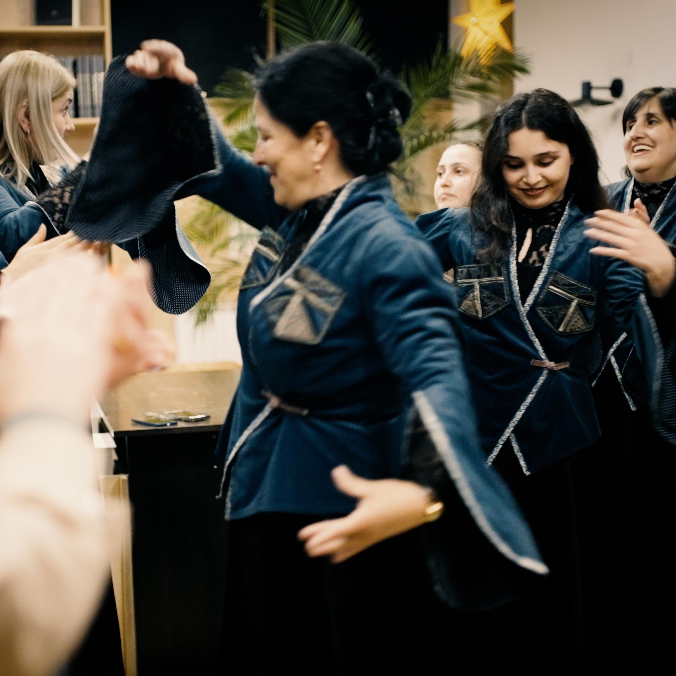 A group of women in matching blue traditional outfits are dancing indoors, with blurred hands clapping in the foreground.