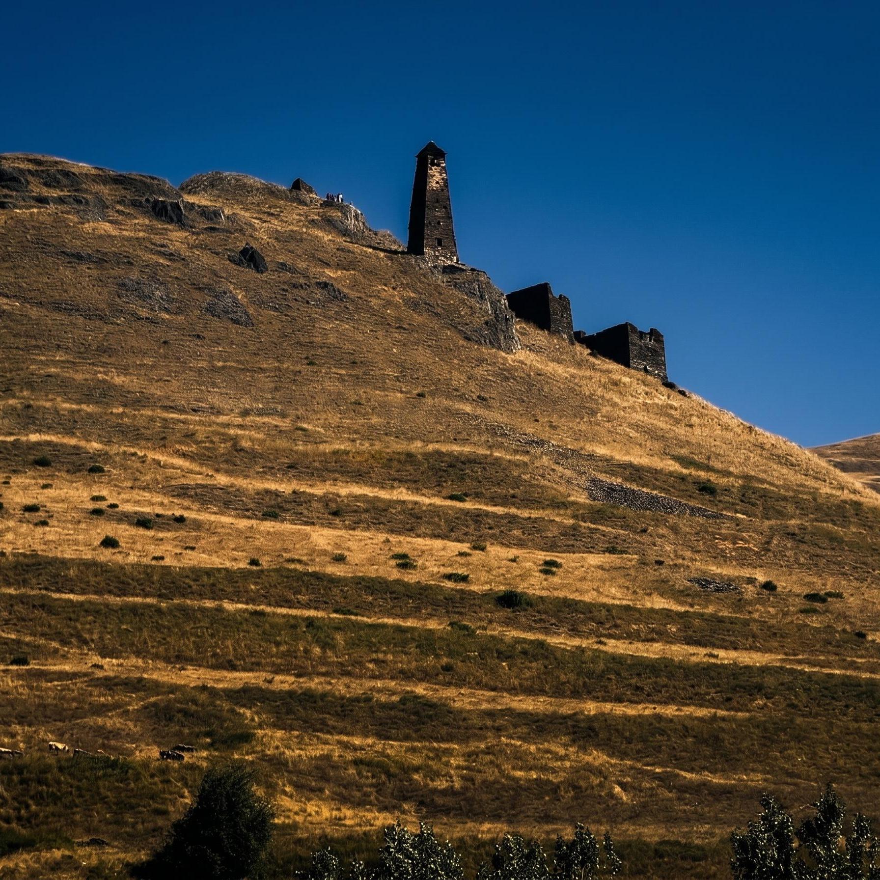 A solitary stone tower and ruins stand on a dry, grassy hillside under a clear blue sky.