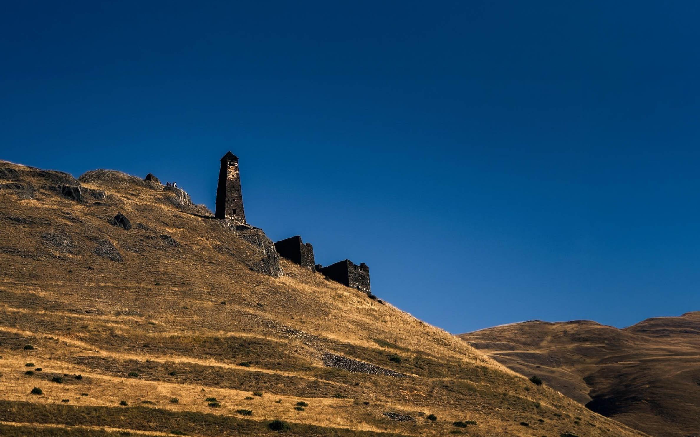 Stone ruins of a tower and walls stand on a dry, grassy hillside under a clear, deep blue sky.